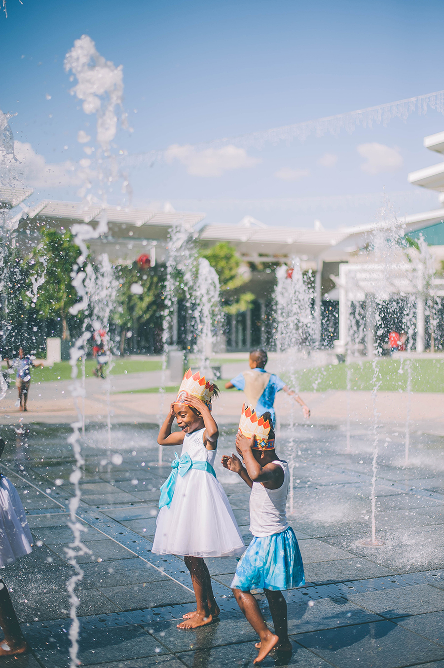 Two children wearing paper crowns play in a water fountain area outdoors on a sunny day, with buildings and more children visible in the background. - Pareto Limited