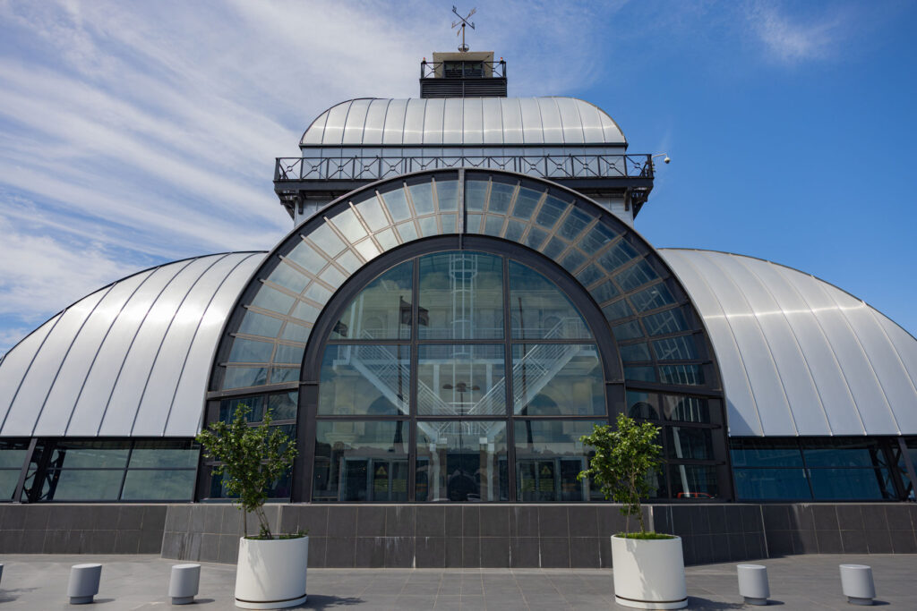 A modern building with a large arched glass entrance, metallic curved roof, and potted plants on a paved area under a blue sky. - Pareto Limited