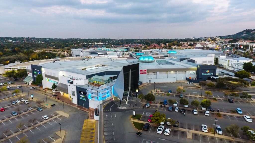 Aerial view of a large shopping centre, part of the Pareto portfolio shopping centres, with a mostly empty car park, modern architecture, and surrounding trees and buildings under a cloudy sky. - Pareto Limited