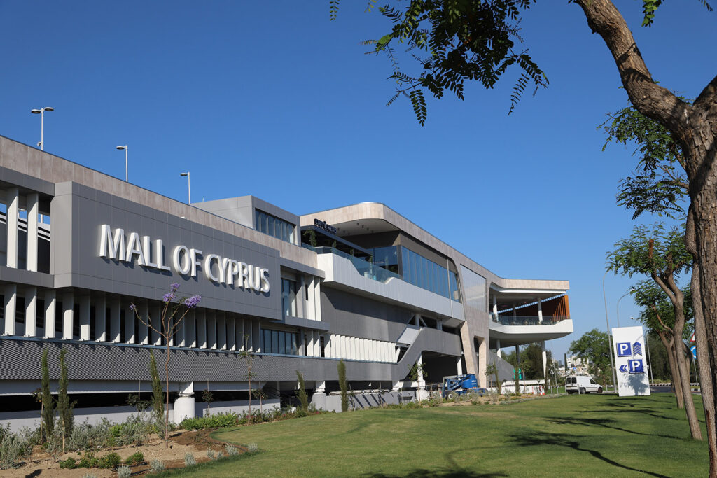 Modern exterior view of Mall of Cyprus, a standout among Pareto portfolio shopping centres, with large signage, green lawn, and parking signs visible under a clear blue sky. - Pareto Limited