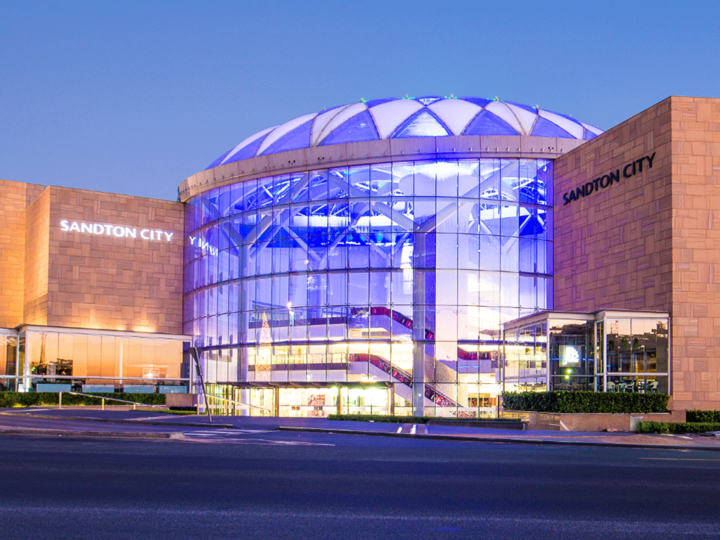 Sandton City shopping centre exterior, part of the distinguished Pareto portfolio shopping centres, features a glass dome, tan stone façade, and illuminated blue and white lights at twilight. - Pareto Limited