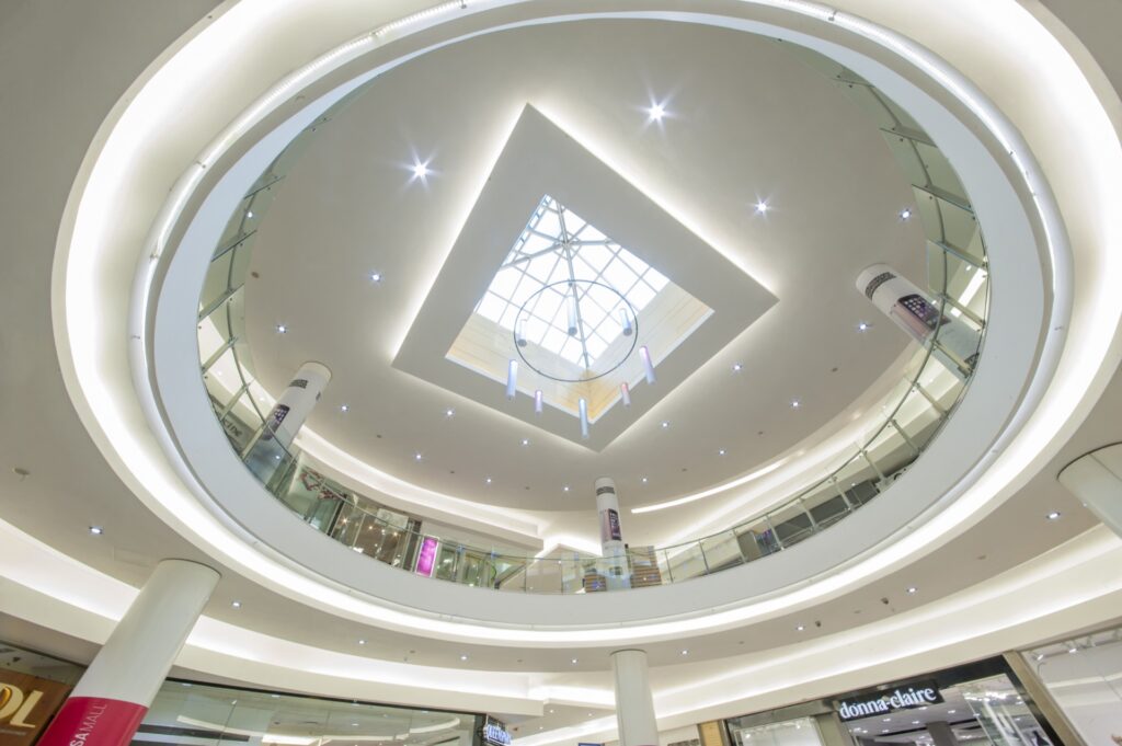 View looking up at Mimosa Mall Bloemfontein’s modern shopping centre circular atrium, featuring glass railings, multiple floors, bright lighting, and a geometric skylight at the ceiling’s centre. - Pareto Limited